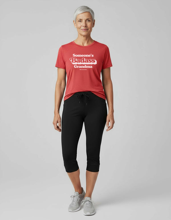 Woman wearing a red t-shirt with Someone's Badass grandma and black pants on a light gray background