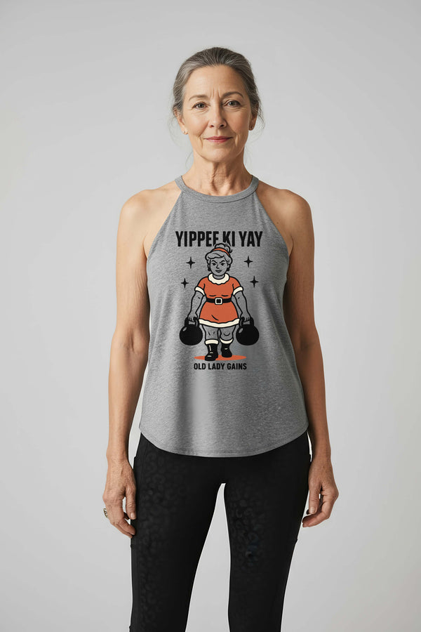Woman wearing a gray tank top with a christmas design that says Yippee Ki Yay and Mrs. Clause doing a farmer carry with two kettlebells on a plain background